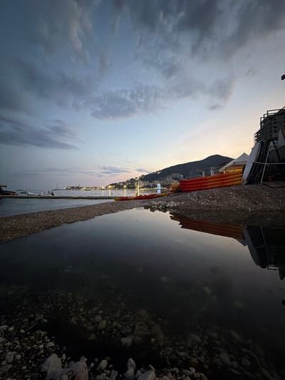The image depicts a waterfront scene in Budva, Montenegro, at dusk. The perspective is rotated 90 degrees clockwise. In the foreground, a rocky embankment lines a body of water. Behind the embankment, several kayaks with orange and red accents are stacked. Further in the background, a town with illuminated buildings stretches along the coastline, nestled at the foot of a hill. The sky is overcast with dramatic clouds, suggesting either approaching or receding stormy weather. The overall mood is serene yet slightly dramatic due to the weather and the twilight lighting. There is no visible text in the image.