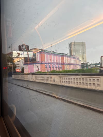 The image captures a view through a rain-streaked window, likely from a moving vehicle, looking out at a city street in Copenhagen, Denmark. In the mid-ground, a brightly colored building with pink and orange stripes is visible. This structure appears to be a commercial building or perhaps a temporary amusement structure, with several windows or openings. Behind it, taller buildings are present, including a modern, light-colored high-rise building. In the foreground, a wet asphalt road and a decorative concrete barrier are seen, suggesting an urban environment during or after rainfall. The sky is overcast, contributing to a subdued lighting condition.