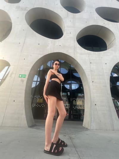 A young woman with dark hair and sunglasses stands in front of a concrete building with large circular openings. She is wearing a black crop top and a black skirt, and black sandals. The building's exterior is rough concrete with several circular cutouts that reveal windows on the interior. The woman is posing with her hands on her hips and her body angled slightly away from the camera. A green sign with "AED" and an arrow is visible on the concrete wall. The scene appears to be outdoors, with bright daylight casting shadows. The overall mood is casual and relaxed, with the woman appearing confident and posed. The architecture is modern and geometric.