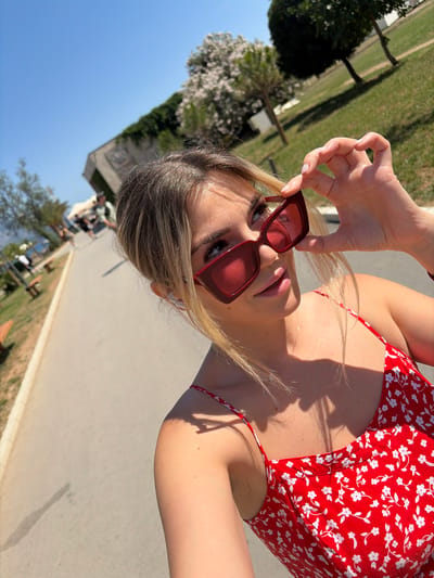 The image shows a young woman with blonde hair wearing red-framed sunglasses and a red floral sundress. She is holding her sunglasses up with her right hand. The woman is outdoors, with a paved walkway and grassy areas visible in the background. There are trees, a building, and a body of water in the distance. People are walking on the walkway in the background. The scene appears to be in Budva, Montenegro, likely on a sunny day.