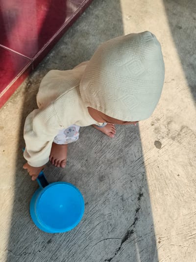 An overhead shot depicts a baby on a concrete surface, wearing a white patterned cap and a long-sleeved, cream-colored sweater with elephant-printed bottoms. The baby's feet are visible, positioned near a blue plastic dipper. A red tiled area is in the upper left corner. The concrete surface is marked with cracks and shadows. The image was taken in Pacet, Indonesia.