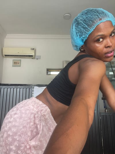 A woman wearing a blue disposable surgical cap, a black crop top, and fuzzy pink sweatpants poses indoors. Her dark skin is visible, and she wears a nose ring and gold hoop earrings. She looks towards the camera with a slight tilt of her head. Behind her is a white wall with an air conditioner unit and a small framed picture. A window and a closed black shutter are also visible in the background.