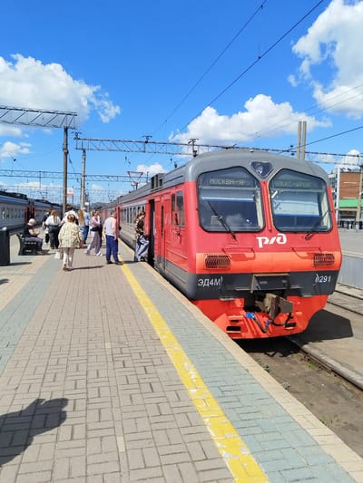 A bright, sunny day at a train station in Moscow, Russia. The sky is a vivid blue, dotted with fluffy white clouds. A red and gray passenger train is parked at the platform, its front clearly visible. The train has "МОСКВА-ЯР" and "АЛЕКС-ДРОВ" displayed on its front windows, indicating its route. The RZD logo is prominent on the front of the train.
Several people are on the platform, some standing and talking, others boarding or disembarking the train. A woman in a plaid shirt and jeans is stepping onto the train, her foot on the lower step. Another person, possibly a man, is standing nearby. Further down the platform, a group of people are gathered, some sitting on a bench, others standing and chatting. A woman in a light-colored jacket is walking towards the train. The platform is paved with interlocking bricks in shades of gray and light blue, with a bright yellow line marking the edge.
The scene suggests a typical day at a bustling train station, with passengers preparing for or concluding their journeys. The sunlight and clear skies indicate pleasant weather, likely mid-day given the length of the shadows. The presence of the RZD logo clearly places the location within Russia.