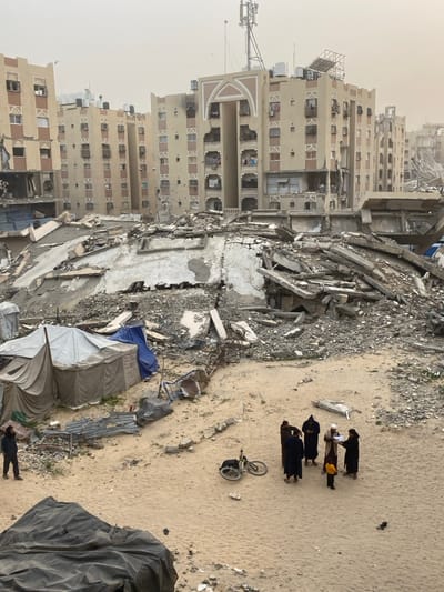 The image depicts a devastated urban landscape in Khan Yunis, Palestinian Territory, under an overcast or hazy sky. In the background, multiple multi-story residential buildings stand, many exhibiting severe structural damage, including partially collapsed sections, blown-out windows, and blackened facades indicating fire damage. Some buildings retain architectural details like arched entryways and balconies, where laundry is visible on less damaged sections. Communication antennas, satellite dishes, and water tanks are present on rooftops.
The midground is dominated by large, irregular piles of concrete rubble, twisted rebar, and debris from collapsed structures, forming a chaotic mound of building remnants. To the left of this rubble, several makeshift tents constructed from light-colored canvas and blue tarpaulins are erected on the sandy ground, suggesting temporary shelter. Scattered metallic debris and broken objects are visible near these tents.
The foreground consists of a relatively open, sandy ground area. On the right, a group of five individuals is gathered. Four adults, dressed in dark, long garments, some with head coverings, surround a smaller child in a yellow top. One adult holds and appears to be reading from a white sheet of paper. A bicycle lies on its side in the sand near this group. To the far left, a lone male figure in dark clothing and a head covering stands facing towards the right. A large, dark tarp or fabric bundle is partially visible in the bottom left corner. The overall scene conveys widespread destruction and makeshift living conditions.