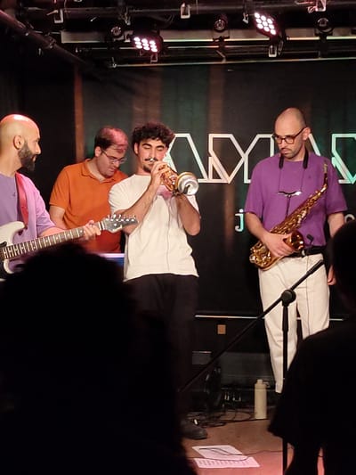The image depicts a live music performance on a stage in Tel Aviv, Israel. Four musicians are visible. On the far left, a bald man with a beard wearing a purple shirt is playing a white electric guitar. Next to him, a man in an orange shirt looks down at his instrument. In the center, a young man with dark, curly hair is playing a trumpet. To his right, a bald man with glasses is wearing a purple shirt and white pants and holding a saxophone. The stage has a black backdrop with a white geometric design. Lighting equipment is visible on the ceiling. The floor is concrete, and music stands and a water bottle are present. In the foreground, the silhouettes of audience members are visible.