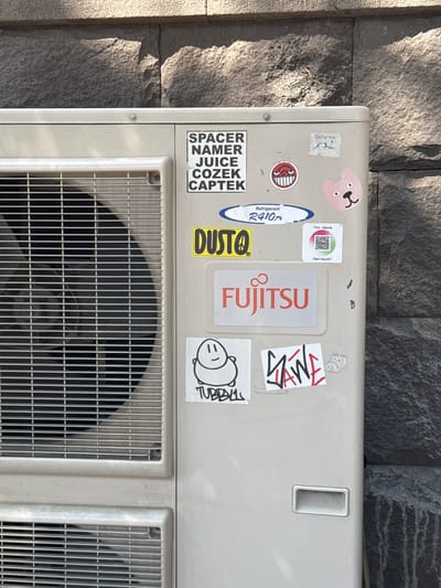 The image shows an outdoor air conditioning unit mounted on a stone wall. The unit is white and has several stickers on its surface. The stickers include brand logos like Fujitsu, a humorous character with a red mouth and wide eyes, and a pink bear face. There are also text-based stickers and a QR code with Russian text. The unit itself appears to be a standard outdoor condenser unit for a split-system air conditioner, with grilles for air intake and exhaust. The stone wall behind it is made of large, rough-hewn grey stones. The lighting suggests it is daytime with sunlight casting shadows. There are no people or animals visible in the image. The scene is likely an exterior wall of a building in a city.