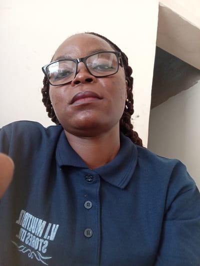 A waist-up, close-up, selfie-style photograph shows a dark-skinned woman with braids and glasses. She is wearing a navy blue polo shirt with a collar and three buttons. The shirt has white lettering on the left side of the chest, which appears to be a logo or text, but it is partially obscured and reversed due to it being a reflection from a mirror. The background is a plain, light-colored wall with a vertical shadow on the right side, suggesting an indoor environment with a light source from the left. The image was taken in Jalingo, Nigeria.