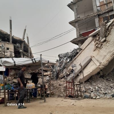 The image depicts a scene in what appears to be Khan Yunis, Palestinian Territory, judging by available context and details visible. A partially collapsed building dominates the background, with debris strewn about. Power lines are visible overhead. In the foreground, a man stands next to a makeshift structure, possibly a market stall. It holds various items, including what looks like packaged goods and textiles. The man seems to be looking or attending to items on the stall. The overall atmosphere suggests destruction and a struggling informal economy.