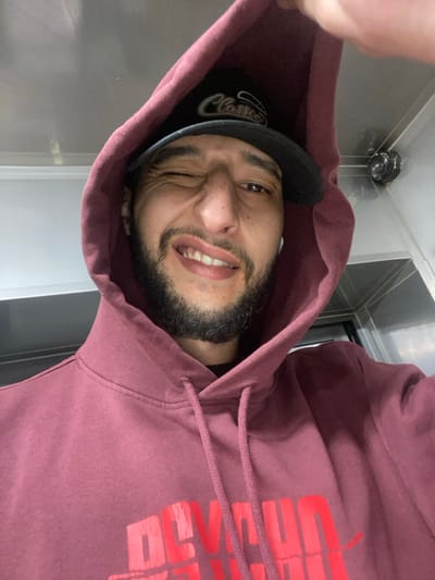 A person wearing a maroon hoodie and black baseball cap with "Classic" embroidered on it is seen from a close, angled perspective. The individual is squinting with a smirk, showing their teeth. A security camera is visible in the background, near a stainless steel-like wall. Part of a red graphic is visible on the hoodie.