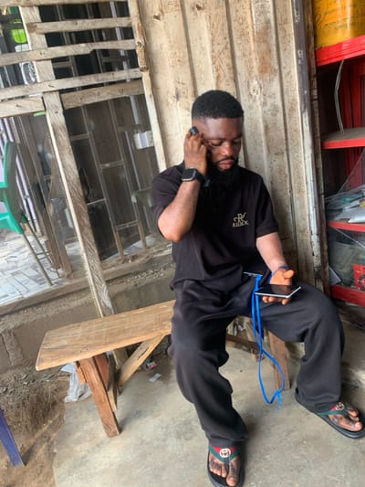 This image depicts a male subject seated on a rustic wooden bench within an urban environment in Okene, Nigeria. The individual, of dark complexion with a short beard and dark hair, is wearing a black short-sleeved collared shirt with a gold "RL REEBOK" logo on the left chest, black trousers, and black slide sandals with green and red striped accents. A black smartwatch is visible on his left wrist.
He is engaged with two mobile phones: one black smartphone is held to his left ear, suggesting an ongoing call or audio interaction, while a second silver-backed smartphone is held in his right hand. A blue cable, likely a charging cable, is connected to the phone in his right hand and extends downwards. His gaze is directed downwards and to his right.
The immediate background to the left features a weathered wooden structure, possibly a window frame or a security grill, with mesh visible and hints of greenery beyond. To the right, vertical wooden planks form a wall, behind which red and yellow shelves hold various items, including a yellow cylindrical container with visible text "Product of Nigeria, Ajao Beach," and a glass display case below. The ground is a concrete slab, appearing worn and somewhat uneven, with dirt and debris visible near the base of the wooden structures. The overall scene suggests an informal commercial setting, such as a shop entrance or a market stall.