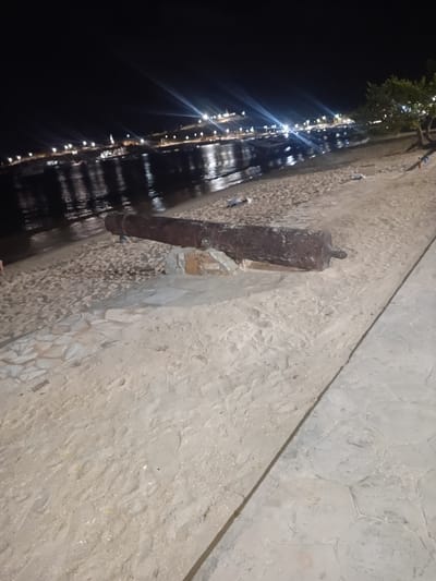 At night, a beachfront scene in Juan Griego, Venezuela, features a sandy area with a large, weathered cannon lying horizontally on the sand. The cannon is positioned on top of a rough stone structure, and its metallic surface appears dark and corroded. The background shows the calm water of a bay or harbor, illuminated by distant city lights and lights from boats. The water reflects the lights, creating streaks on its surface. On the sandy shore, there are scattered pieces of debris and some footprints. A paved walkway with irregularly shaped stones runs along the right side of the image, separating the sand from a darker area. A tree with green foliage is visible in the upper right corner of the frame. The overall atmosphere is dark, with artificial lights providing the primary illumination.