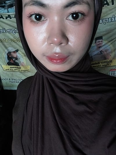 This is a close-up, vertical, selfie-style photograph of a young woman wearing a dark brown hijab. The lighting is warm and appears to be artificial, creating a slight glow on her skin. Her face is tilted upwards, and her eyes are looking directly at the camera. She has noticeable makeup, including defined eyelashes, blush, and a glossy, bright red lipstick that gives her lips a plump appearance. The background is a textured surface with some text visible, including "DEZEMBEL" and what appears to be Indonesian text. The text also includes a year, "2003", and some Cyrillic script, which is unusual given the context. There are also images of people in traditional attire in the background. The scene appears to be indoors, possibly in a domestic setting. The overall impression is one of youthful vanity and a casual, personal moment captured for social media. The combination of the woman's makeup, the specific text elements, and the Indonesian location context creates a unique and somewhat eclectic visual.