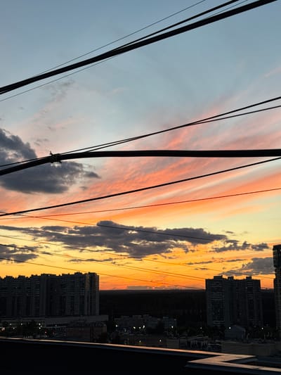 This media file captures a sunset over Khimki, Russia, viewed from a high vantage point. The scene is dominated by a dramatic sky painted with vibrant hues of orange, pink, and yellow, streaked with dramatic clouds. Several power lines are prominently featured in the foreground, stretching diagonally across the frame and obscuring parts of the sky and cityscape.

Below the colorful sky, a cityscape unfolds, characterized by numerous tall apartment buildings that are silhouetted against the setting sun. The buildings are densely packed, suggesting an urban environment. Some windows in the buildings are lit, indicating evening activity. The overall atmosphere evokes a sense of calm and the beauty of a fading day in an urban setting. The time of day is clearly evening, as evidenced by the sunset and the lit windows. The weather appears clear, with clouds contributing to the spectacle of the sunset. The location is confirmed as Khimki, Russia. There is no visible text in the media file.