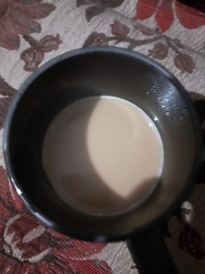 The image captures a top-down view of a dark-colored mug or small saucepan filled with a light brown, creamy liquid, most likely tea or coffee with milk. The container appears to be made of a dark material, possibly metal, and has a subtle handle visible extending to the right. The surface of the liquid is smooth and shows a distinct curved reflection, indicating a light source from above and slightly to the left. The container is placed on a patterned fabric surface, which serves as the background. This fabric features repeating floral or leaf motifs in shades of maroon or deep red, intermingled with lighter brown or beige tones, against a lighter, possibly off-white or light brown base. The overall lighting suggests an indoor setting. No people or text are visible in the image.