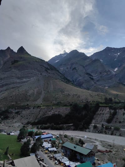 An aerial perspective captures a sprawling village nestled beside a meandering river, dwarfed by majestic, snow-capped mountains under a cloudy sky in Gushal, India. The village, a cluster of buildings with predominantly blue and green roofs, lies on the lower right and center of the frame, bordered by a road and a lush green field on the lower left. Vehicles, including cars, trucks, and vans, are parked along the road and in designated areas. The mountainous landscape dominates the upper portion of the image, showcasing rugged terrain with sparse vegetation and visible snow on the peaks. The river, a murky brown, flows through the valley, reflecting the overcast sky.