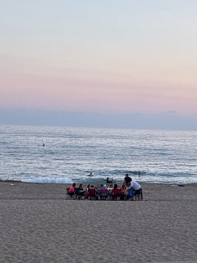 A group of people are gathered on a sandy beach, facing the ocean. The sky is a gradient of pale blue to pink, indicating either sunrise or sunset. Several individuals are seated in portable chairs around a low table, some appear to be talking or interacting. One person, wearing a white t-shirt and dark shorts, stands near the seated group, possibly engaged in conversation or handing something to another person. In the ocean, several people are visible, some swimming or wading in the water, and at least two individuals are on surfboards further out. The waves are moderate, with white foam crashing on the shore. The sand appears textured, with visible footprints or patterns. The overall scene suggests a relaxed gathering on the beach. This scene is consistent with the beaches in the city of Alanya, Turkey, known for its coastal location on the Mediterranean Sea.