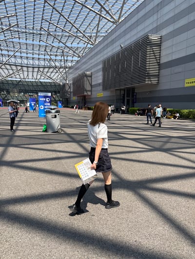 A woman in a white shirt, dark shorts, black socks, and black shoes stands on a concrete surface with shadows from a structural framework. She holds a yellow document with circular graphics. The location is outside a large building with a glass roof structure and metal facade. Blue signs are visible with directional information. Other people are present in the background. This scene is located in the city of Liangjiang, China.
