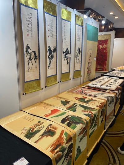 The image depicts a display of traditional Chinese scroll paintings, likely at an art exhibition or market. The scrolls are hung vertically and are presented in a row, suggesting a curated collection. The subjects of the paintings vary, with visible depictions of horses in dynamic poses, possibly in mid-gallop, and landscapes featuring mountains and foliage. One scroll appears to feature a figure, possibly a deity or a serene individual, in a flowing robe. The background suggests an indoor setting with spotlights illuminating the artwork. The scrolls themselves are rendered on what appears to be rice paper, some with ornate golden borders and dark rollers at the top and bottom. The overall presentation is neat and organized, with the scrolls unfurled to showcase the artwork. A small card with text and an image is visible on the bottom right, likely providing information about the artwork or artist. The setting appears to be well-lit, indicating it's likely daytime or a well-lit indoor venue. The presence of multiple scrolls and the way they are displayed points to an event focused on the appreciation or sale of Chinese art. The visible text, though partially legible, is in Chinese characters, common for such artworks.
