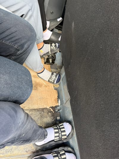 The image shows the feet of three individuals inside what appears to be a vehicle. The flooring of the vehicle is covered with torn cardboard and some metal components are visible. One person is wearing white socks and black slides with white stripes and the words "ENJOY LIFE" printed on them. Another person is wearing grey socks with the word "Super" printed on black slides. A third person is wearing white socks and black slides. The interior of the vehicle is dark and appears to be utilitarian. The scene is a close-up, low-angle shot focusing on the feet and lower legs of the individuals. No specific details about the city of Janowice, Poland, are discernible from the image.