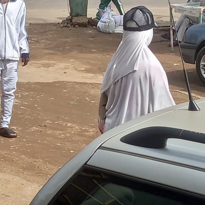The image depicts a scene in what appears to be a public area in Dadi Riba, Nigeria. A person is fully covered in a white sheet, with only a black mesh cap visible on their head. They stand next to a parked white vehicle, with a portion of the car door and handle visible. In the lower foreground, the legs and torso of another person wearing a white tracksuit with blue stripes are visible. The ground appears to be a mix of dirt and paved surface. In the background, there are hints of greenery and other parked vehicles.