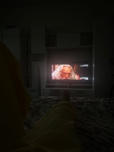 In a dimly lit bedroom in Shushary, Russia, a person lies on a bed covered with a patterned comforter, facing a television screen. The person's legs, covered in yellow pajamas, are visible on the bed, with one foot extending towards the television. The television displays a close-up of an elderly man with white hair and glasses, likely a character from a movie or show. Text overlays are visible on the television screen, suggesting content related to subscription services or channel information. The room contains a white wardrobe unit behind the television, with shelves and cabinets. A tall, white standing fan is positioned between the bed and the wardrobe. The overall atmosphere is one of relaxed, solitary viewing in a domestic setting.