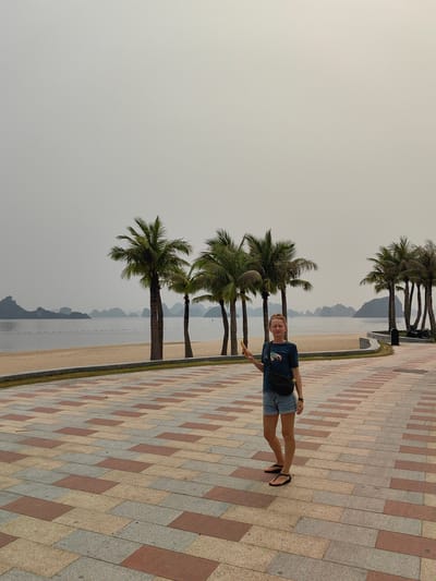 The image features a woman standing on a patterned paved walkway alongside a sandy beach and bay, characteristic of Ha Long Ward, Vietnam. The woman, positioned slightly left of center, faces the camera with her eyes closed or squinting and a gentle smile, holding a yellow smartphone in her right hand. She wears a dark blue t-shirt with a graphic design, light denim shorts, and black flip-flops, with a black fanny pack worn across her chest.
Behind her, several tall palm trees line the edge of the sandy beach, which borders a calm body of water. In the distance, numerous iconic karst islands or limestone formations rise from the bay, partially obscured by an overcast and hazy sky, which is a uniform light grey. A line of buoys or markers stretches across the water.
The weather appears hazy and diffused, creating soft, muted lighting. The paved walkway beneath her feet is adorned with a mosaic pattern of rectangular tiles in various shades of red, beige, and grey. The overall scene conveys a tranquil, relaxed atmosphere in a coastal setting during daytime, despite the overcast conditions. No explicit text is visible.