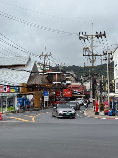 This is a vertical, eye-level shot of a street in Karon, Thailand, on an overcast day. The foreground is dominated by a grey asphalt road with yellow lane markings. Several cars and motorcycles are parked or moving along the road. To the left of the road, a dense network of utility poles and wires stretches upwards, creating a chaotic visual. Adjacent to the poles are buildings with shops and signs, featuring a mix of Thai and English text, including "SAMSUNG," "HUAWEI," "realme," "vivo," "oppo," "ART," "AIS," "KIRI," and "EXCHANGE." One license plate reads "ณบ 5498". A person in a blue shirt and white shorts is visible near a blue industrial-looking machine in front of one of the shops. In the background, a lush green, tree-covered hill rises under the cloudy sky. The overall impression is a busy, somewhat cluttered urban street scene in a tropical location.