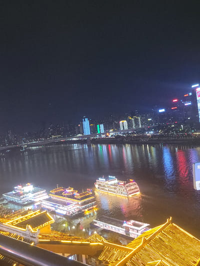 The image captures a vibrant night cityscape along a wide river, likely in Yuzhong District, China. The scene is dominated by extensive artificial illumination from buildings and boats, creating strong reflections on the water.
On the far bank of the river, a dense skyline of modern high-rise buildings is brightly lit with various colors, including red, blue, green, and white. These lights cast long, colorful streaks across the dark river surface. A bridge, also adorned with lights, spans the river in the distance.
The river itself is a central feature, teeming with activity. Several illuminated boats, likely cruise ships, navigate the waters, showcasing their own elaborate lighting patterns, primarily in white and red. Their movement creates gentle ripples that slightly distort the reflections of the city lights.
On the near bank, a complex of multi-tiered buildings, characterized by traditional Chinese architectural styles, glows with a striking golden-yellow light. These intricate structures extend along the waterfront, suggesting a prominent cultural or tourist area. A metal railing is visible in the bottom right foreground, indicating the photograph was taken from an elevated vantage point, possibly a balcony or a viewing platform.
The time of day is clearly night, with the sky appearing dark and generally clear. The extensive and diverse artificial lighting, from the towering skyscrapers to the traditional facades and river boats, imbues the scene with a dynamic and festive atmosphere. Some Chinese characters are visible on distant buildings, including a clear "南" (nán) on a blue sign and hints of "古" (gǔ) on a building on the near side, though most text is too small or blurred for precise identification. The overall impression is a bustling and visually rich nocturnal urban environment.