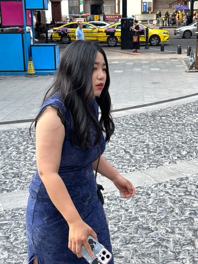 A medium shot shows a woman with long, dark hair, wearing a blue dress and holding a cell phone with a cartoon design. She is standing on a sidewalk in Yuzhong District, China. To the left of the woman, there are taxis and various blue and pink structures, possibly booths or displays. The sidewalk is gray and appears to be made of stone or textured concrete.