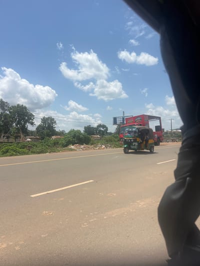 The image depicts a street scene in Rafin Par, Nigeria, on a sunny day with scattered clouds. In the foreground, a paved road with a yellow dividing line is visible. A three-wheeled auto-rickshaw, painted green and yellow, is traveling on the road, displaying the license plate "PTA 10043". Behind the auto-rickshaw is a large red truck. The roadside on the left is lined with lush green trees and vegetation. Further back, buildings and more trees can be seen. A significant amount of refuse is piled along the side of the road, creating a noticeable environmental issue. The sky is a clear blue with fluffy white clouds. The perspective suggests the photo was taken from inside a vehicle.