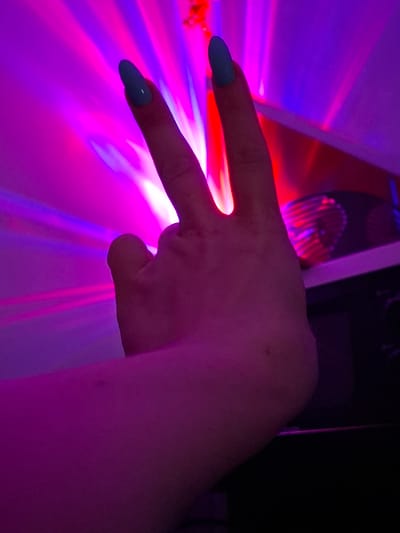 An upper body shot of a person's left hand is visible, with their index and middle fingers extended upward and slightly apart, forming a 'V' shape. The fingernails are painted a light blue. The hand is illuminated by vibrant pink and purple light, suggesting a dimly lit environment with colored lighting, possibly from a disco ball or stage lighting. The background features streaks of light radiating outwards and a dark object with a circular, ribbed surface on the right. The scene is devoid of any discernible urban context or landmarks of Zemun Urban Municipality, Serbia. The focus is on the hand and the dramatic lighting.