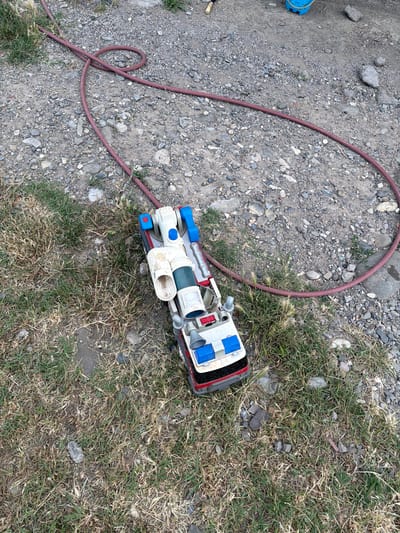 A toy vehicle, resembling a futuristic ambulance or rescue vehicle, lies on a patch of dry, overgrown grass and gravel. The toy is primarily white and blue, with red accents and what appear to be silver detailing around some of its components, such as a large cylindrical tube and smaller nozzles. It has a boxy, segmented body, with various protruding parts and what could be wheels or tracks visible on the sides.

A red garden hose is coiled and draped across the gravelly area, with one end disappearing out of frame and the other lying near the toy. The ground consists of loose, grey gravel mixed with sparse patches of dry, light-green grass and small, grey rocks. A small blue object, possibly another toy or part of equipment, is visible in the upper left corner of the image.

The scene appears to be outdoors, under daylight conditions. The dry grass and gravel suggest a somewhat arid or unkempt environment. The overhead perspective of the image allows for a clear view of the toy and its surroundings. No people are visible. There is no discernible text on the toy vehicle. The location context of Tbilisi, Georgia, hints at a possible urban or suburban outdoor setting. The overall impression is one of discarded or forgotten playthings in a naturalistic outdoor environment.