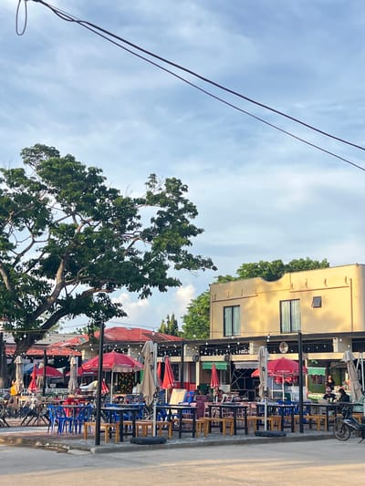 The image depicts an outdoor seating area at a restaurant or cafe in Cuyo, Philippines. The scene is set on a paved surface, likely a street or plaza, with an overcast sky overhead.
In the foreground, numerous tables and chairs are arranged in a scattered fashion, suggesting a casual dining environment. Many of the tables are occupied by chairs of various colors, including blue, red, and white. Several red umbrellas, some open and some closed, are positioned over the tables, providing shade.
A large, leafy tree dominates the left side of the frame, its branches extending over a portion of the seating area. Beyond the tables, a building with a yellow facade and dark-framed windows is visible. A covered walkway or patio runs along the front of this building, and more tables and chairs are set up beneath it. String lights are suspended along this walkway.
In the background, additional structures with red roofs are present. Some people are visible in the distance, including a person walking near the tree on the left and individuals seated at tables in front of the yellow building. A motorcycle is parked on the right side of the image. The overall impression is a vibrant, communal outdoor space, typical of a local establishment in the Philippines.