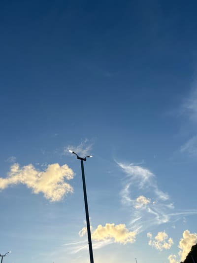 The image shows a clear blue sky with scattered white and yellow clouds. A tall street lamp with two lights is prominently featured in the center-right of the frame, silhouetted against the sky. There are glimpses of other street lights in the lower left and lower right corners. The scene appears to be an outdoor urban environment in Ratsada, Thailand, possibly a street or public area. The lighting suggests it is either early morning or late afternoon. No people or actions are visible.