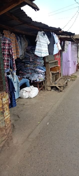 The image captures an outdoor market stall situated in Nairobi, Kenya. The primary subject is a semi-enclosed structure built from rough wooden beams and corrugated iron sheets, some of which are painted purple. Underneath the corrugated roof, numerous articles of clothing, predominantly shirts in various patterns and colors (including plaid and striped designs), are displayed on hangers. Behind these hanging clothes, large piles of folded garments are stacked high. A single individual, identified by their legs and lower torso clad in blue jeans, is seated on a small wooden stool amidst the merchandise, partially obscured by the piles of clothes. A large white, irregularly shaped sack or bundle rests on the ground beside the seated person. To the right, within the stall, there is a simple wooden shelving unit and a low wooden bench-like structure. The foreground consists of a worn, dark grey asphalt or paved ground surface. The upper right portion of the image reveals a clear blue sky, with faint power lines visible.