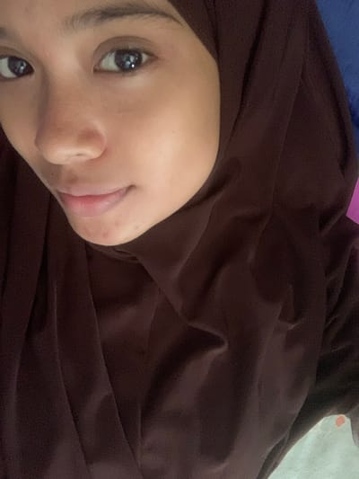 A close-up, self-portrait image depicts a young woman's face, partially covered by a dark brown hijab. The focus is on her eyes, nose, and mouth. Her dark eyes are looking directly at the camera. The skin on her face appears to have some minor blemishes. The background is blurred, with a hint of blue and pink on the right side, suggesting an indoor setting. The image was taken in Sungai Meriam, Indonesia.