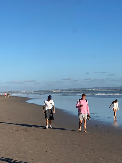 This image depicts a view of Kuta Beach in Kuta, Indonesia, under a clear blue sky with scattered clouds. The sandy beach stretches into the distance, with gentle waves lapping at the shore. In the foreground, three individuals are walking along the wet sand. A man with tattoos on his arms and legs, wearing a white t-shirt and dark shorts, walks towards the camera. To his right, another man in a bright pink shirt and grey shorts is looking at his phone while carrying a pair of shoes in his hand. Further in the distance, a woman in a white dress is walking away from the camera. Other beachgoers can be seen scattered along the shoreline and in the shallow water, with buildings and the coastline visible in the background. The lighting suggests it is late afternoon or early morning.