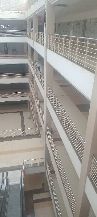 The image depicts the interior of a multi-story commercial or public building located in Kisumu, Kenya, likely an atrium or mall. The scene is observed from an elevated perspective, looking downwards and across multiple levels.

The building features a modern architectural design with light-colored walls, pillars, and floor tiles in shades of beige, off-white, and light grey. The floor pattern consists of large square or rectangular tiles with contrasting lighter and darker geometric outlines. Each floor is defined by horizontal walkways equipped with metallic railings composed of multiple horizontal bars, providing a consistent safety barrier across all visible levels.

At least two individuals are present. One person, wearing a light-colored shirt and dark trousers, is visible on an upper floor, leaning over a railing on the left side of the frame, looking down into the open space. A second, partially obscured individual in a light-colored shirt is visible further up on another floor, also near a railing. No discernible interactions between them are visible.

In the lower-left corner of the frame, a set of downwards-moving escalators is present, indicating access to a lower floor or basement level. Overhead, a roof structure with translucent panels or open sections suggests natural light penetration. A red rectangular sign or object is affixed to a wall on an upper level, though its details are illegible. The environment appears clean and well-maintained, with minimal pedestrian traffic observed at the time of the photograph.
