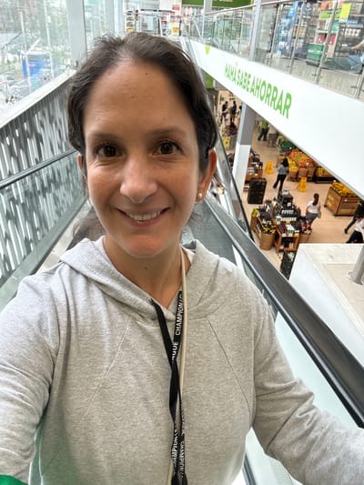 A selfie shot depicts a woman with dark hair and a gray hooded sweatshirt standing on an escalator in a supermarket in Miraflores, Peru. She is smiling at the camera.

Below the escalator, the supermarket aisle is visible. Shoppers are browsing displays of fruits, vegetables, and bottles of wine. A sign reading "MAMA SABE AHORRAR" in green letters is visible above the aisle. A glass barrier surrounds the escalator.