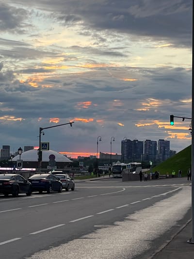 This is a vertically oriented, medium shot image of a street scene at sunset. The sky is filled with dramatic clouds, tinged with orange and yellow from the setting sun, contrasting with darker gray clouds. In the distance, tall modern buildings create a city skyline.

The foreground shows a paved road with white lane markings. Several cars are parked along the right side of the road. A sidewalk or pedestrian area runs alongside the road, with a few people visible walking and standing. Lampposts and traffic signals are present. A traffic light shows a green signal. Various street signs are visible, including a "STOP" sign with Russian text.

The scene suggests an urban environment, possibly near a park or green space on the left side of the image, which is covered in grass. The overall atmosphere is serene, with the vibrant sunset colors contributing to a sense of calm and beauty. The time of day is clearly sunset, indicated by the lighting and sky colors. The presence of cars and people suggests that this is a public area where daily activities are taking place.