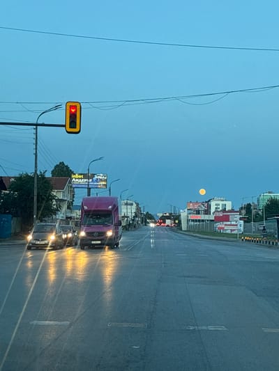 Here is a description of the image:

The image depicts a street scene in Sofia, Bulgaria, likely taken during dawn or dusk. A straight, wet road stretches into the distance, reflecting car headlights. A bright moon hangs in the sky above distant buildings. Traffic lights show a red light on the left side of the frame. A van and other cars are visible on the road. A billboard advertising "inbet" is next to the road. The license plates on the vehicles bear Bulgarian numbering (PK 3808 Bx, CB 9681 CO), indicating the location as Bulgaria. Another billboard is advertising "SELECT" with the words "SELECT" stacked on top of each other.