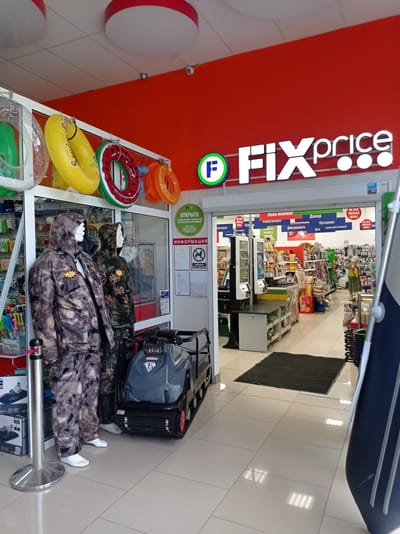 The image shows the entrance of a Fix Price store in Degtyarsk, Russia. The store is decorated with inflatable rings hanging from the ceiling. Inside, there are mannequins dressed in camouflage clothing, a snowmobile, and shelves stocked with various items. The store's signage is visible, with the Fix Price logo prominently displayed. The lighting suggests it is daytime, and the overall atmosphere is that of a retail environment.