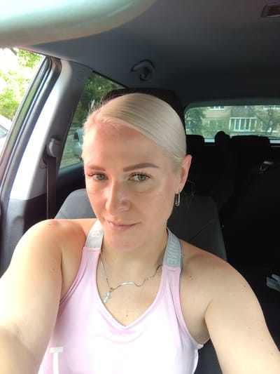 A young Caucasian woman with blonde hair pulled back, green eyes, and light skin is pictured from the chest up, sitting in the driver's seat of a car. She is wearing a pink tank top with grey straps and a silver necklace with a cross pendant. Her left ear has a silver safety pin earring. The background of the car interior shows black car seats and windows through which trees and a building are visible. The setting appears to be a residential street in Kyiv, Ukraine, with the natural light suggesting it is daytime.