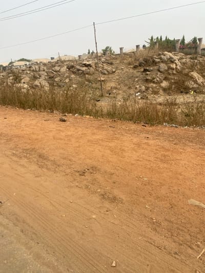 A landscape photograph shows a dirt road curving to the right, bordered by a rocky hillside to the left. The hillside is covered in dry grass and scattered rocks, with some buildings and trees visible in the background. Power lines stretch across the sky on the left side of the image. The dirt road is a reddish-brown color, and tire tracks are visible in the dirt. The sky is overcast.