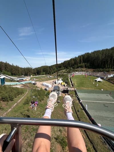 A point-of-view shot from a ski lift looking down on a grassy area, likely a sports complex, possibly for ski jumping. The perspective is from high above, with the ski lift cable visible to the left. A forest lines the top of the frame. The sky is blue with light clouds. Legs with white socks and sneakers are on the right side of the frame. The ski lift's metal bar is visible. In the background are people, tents, and what appears to be a ski jump ramp. The location is in Прикамский, Russia.