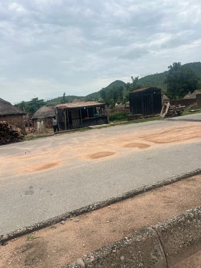 The image, taken in Kona Garu, Nigeria, depicts a roadside scene with several structures and a person. The viewpoint is from a slightly elevated position looking downwards and to the right. The sky is overcast with pale grey clouds.
In the foreground, a paved road runs diagonally from the lower left to the upper right of the frame. The road surface is asphalt, with some lighter brown, sandy patches and a distinct dark stain. To the right of the road is a gravelly shoulder, separated by a rough, rocky edge.
Along the left side of the road, a collection of buildings and structures is visible. Some appear to be made of wood and corrugated metal, with weathered, rusty roofs. One larger structure has dark, vertical metal siding. There are also thatched-roof buildings and piles of firewood. A person wearing a light-colored shirt and dark pants is standing near one of the structures, looking towards the road. Scattered around are other objects, including what appears to be plastic sheeting and possibly some agricultural produce.
The background features a lush, green hillside covered in trees and vegetation, suggesting a rural or semi-rural environment. The overall impression is of a functional, perhaps somewhat rudimentary, community living alongside a main thoroughfare. The time of day is likely daytime, given the ambient light, and the weather is cloudy. No text is visible in the image.
