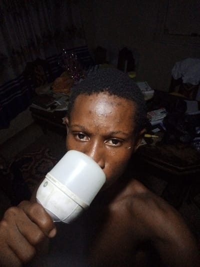 A close-up of a person with dark skin drinking from a white plastic container. The person is looking directly at the camera. The background is dimly lit and cluttered with various objects, including furniture and fabrics. The image appears to be taken indoors. The location is stated to be Ukwu-Abwa, Nigeria.