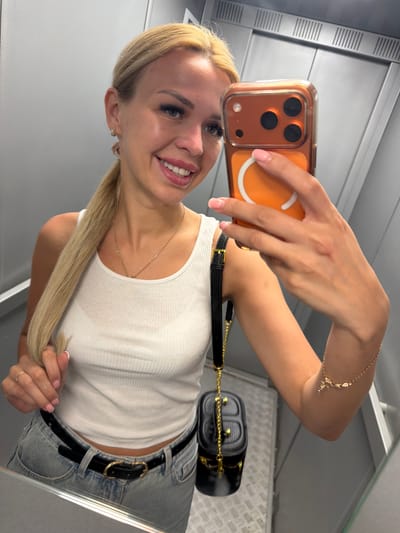 A young woman with blonde hair pulled back in a ponytail, wearing a white tank top and blue jeans, takes a selfie in an elevator. She has on a gold necklace and earrings, a black belt with a gold buckle, and a small black handbag with a gold chain strap is slung over her shoulder. She is holding an orange-cased smartphone in her right hand. The elevator is dimly lit, with grey walls and a metal floor. The scene is indoors.