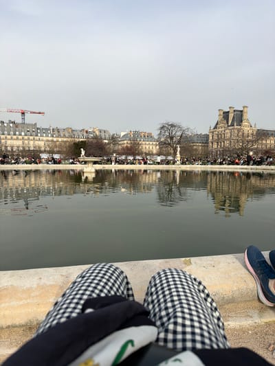 The image depicts a scene from the Jardin des Tuileries in Paris, France, specifically from the edge of the Grand Bassin Rond. The perspective is low-angle, from the viewpoint of a seated individual whose lower legs, clad in black and white gingham pants, occupy the immediate foreground. A dark garment rests on the lap, partially obscuring a white garment with green and gold detailing. To the right, a pair of dark blue and pink athletic shoes are positioned on the light-colored concrete coping of the basin.
The midground consists of the still, dark grey-green water of the large circular basin, reflecting background structures and bare trees. Beyond the basin, numerous individuals are visible, seated on green park chairs or standing, populating the park grounds along the distant edge. The background skyline features a continuous line of classic Parisian Haussmannian buildings with grey zinc roofs on the left and center, above which a red and white construction crane is prominently visible. Interspersed among bare deciduous trees are white statues and a central fountain. On the far right, a large, ornate building with multiple distinct chimneys and a dark mansard roof, consistent with the architecture of the Louvre Palace, stands against the uniformly overcast light grey sky.