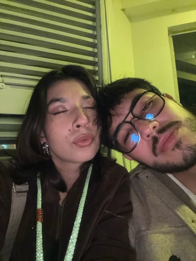 A man and a woman are closely posed together. The man has a beard and wears glasses. The woman has her eyes closed and is wearing a brown jacket with a sparkly lanyard. They appear to be indoors, against a background with vertical lines suggesting window blinds or a similar structure. The overall lighting gives the photo a slightly yellow tint.